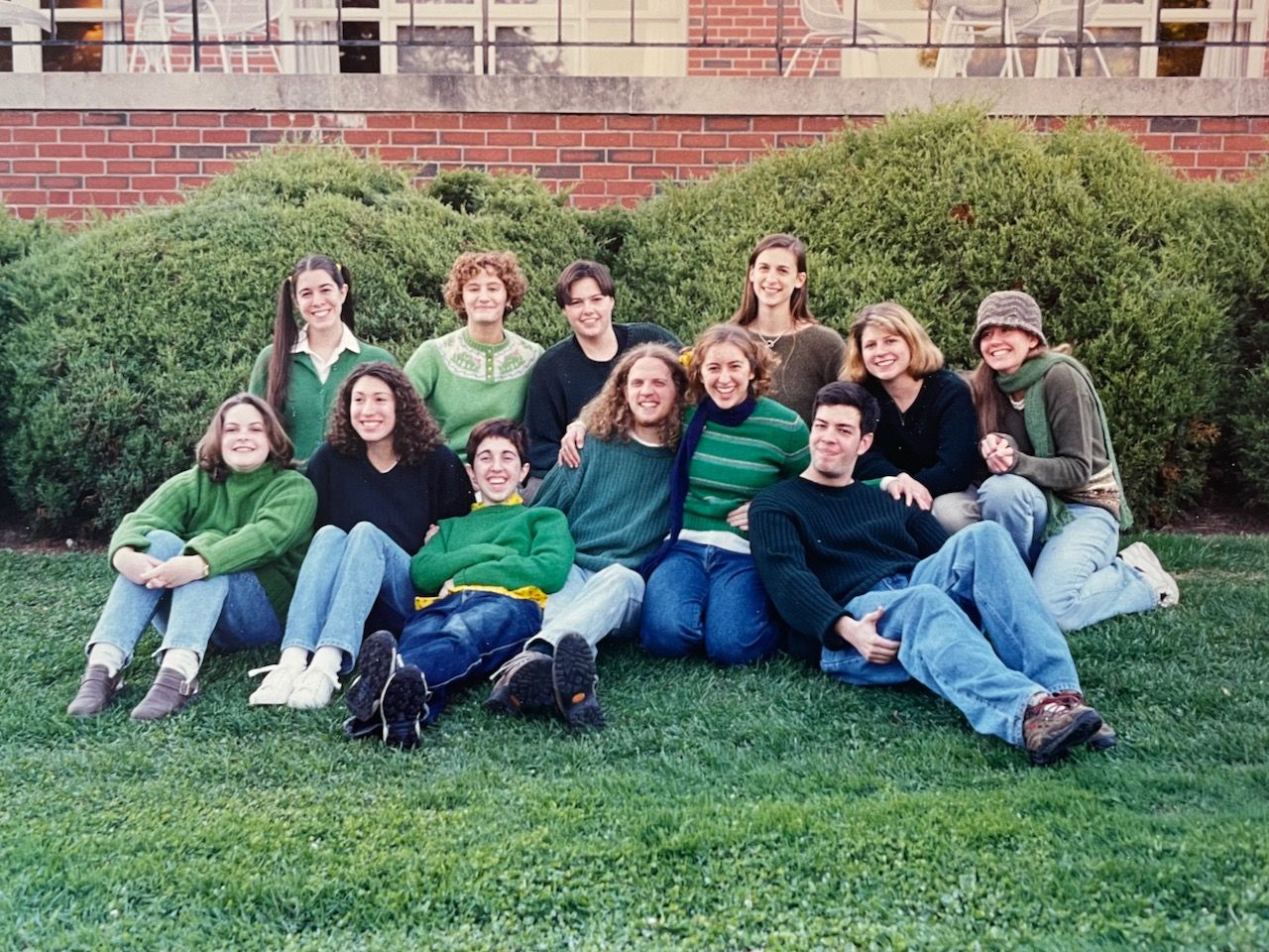 Group of 12 smiling college students in green sweaters, on a green lawn with green shrubs behind them.